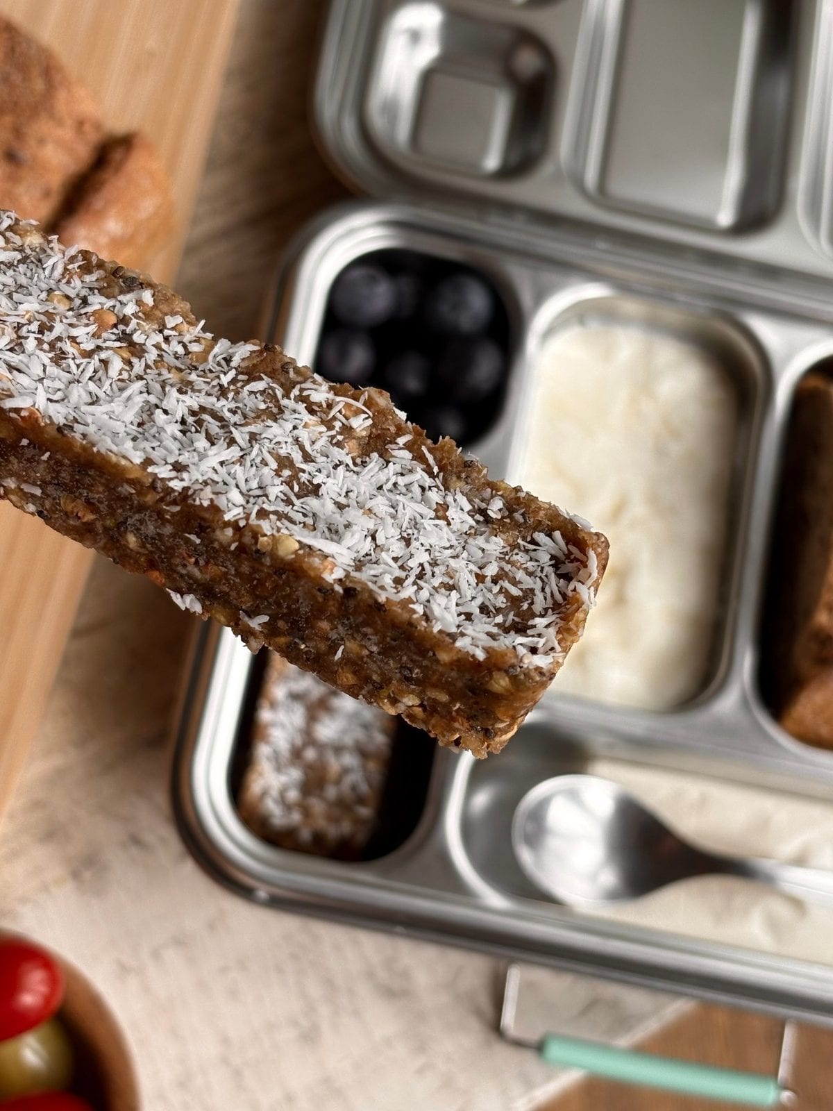 A hand holding a chewy frozen coconut lemon bar topped with shredded coconut over a stainless steel bento lunchbox with blueberries, yogurt, and more bars.