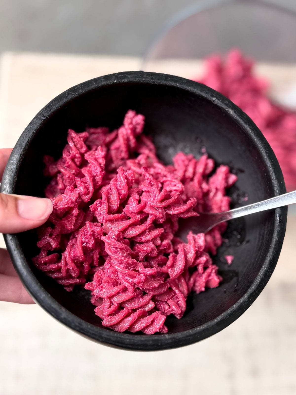 A black bowl of bright pink fusilli pasta coated in a creamy beetroot and tofu sauce, held in one hand with a fork.