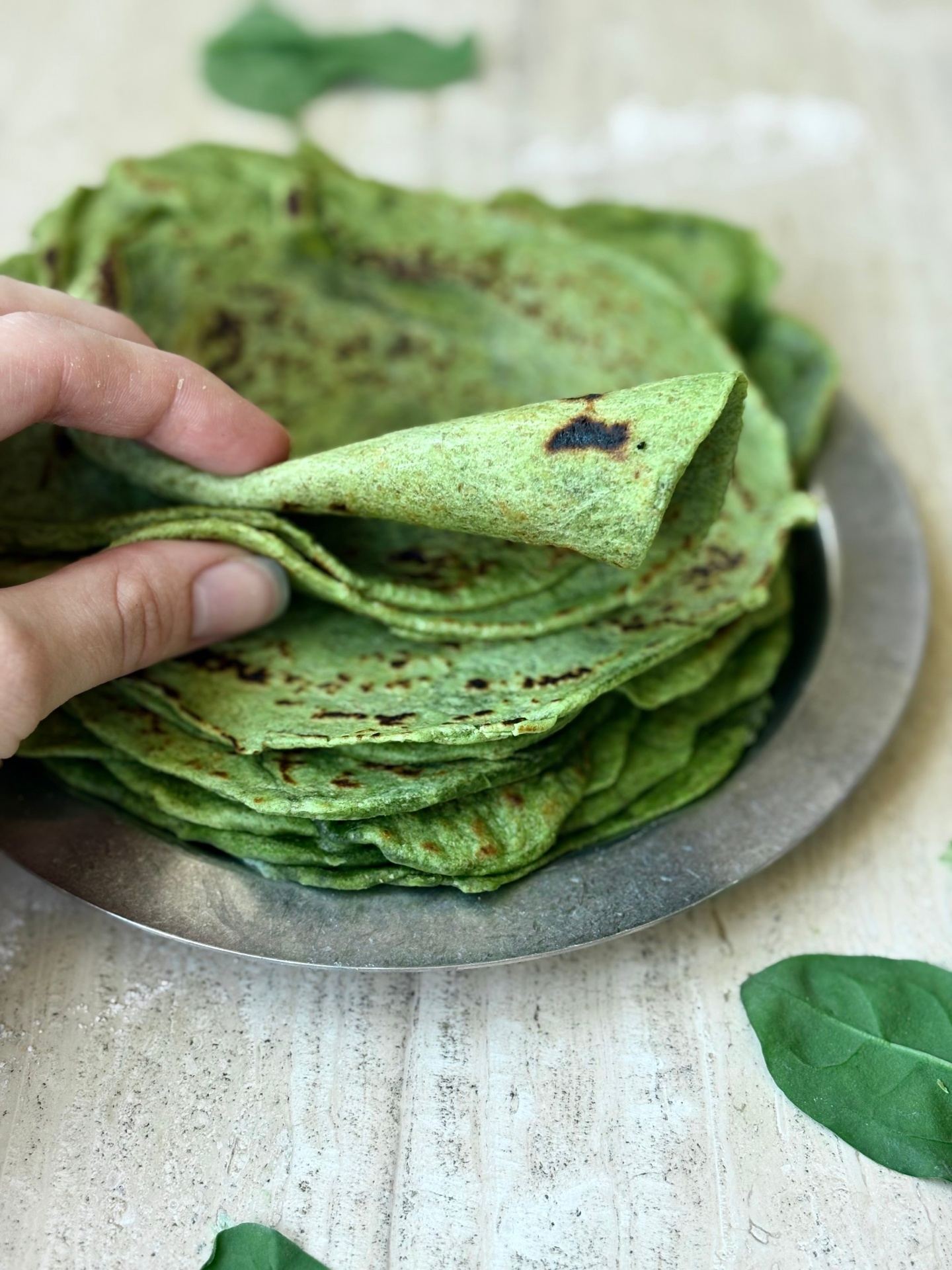 A hand lifting a soft, rolled-up green spinach wrap from a stack of freshly cooked wraps on a metal plate, with fresh baby spinach leaves scattered around.